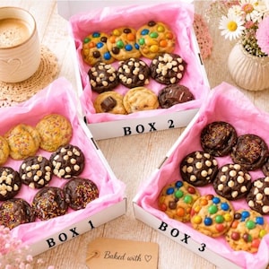May include: Three white cardboard boxes labelled BOX 1, BOX 2, and BOX 3, each lined with pink tissue paper and filled with an assortment of baked cookies. The cookies include varieties with colourful candy pieces, white chocolate chips, and sprinkles.