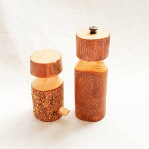 May include: Two wooden salt and pepper shakers made from tree bark. The shakers are cylindrical and have a natural wood finish. The pepper shaker has a small metal knob on top.