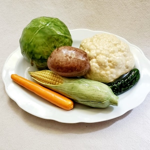 May include: A white ceramic plate with a variety of vegetables, including a green cabbage, a brown potato, a white cauliflower, a green corn on the cob, a green cucumber, and an orange carrot.