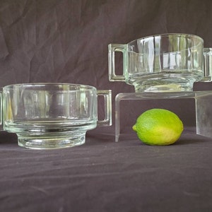 May include: Two clear glass bowls with handles, one on top of a clear acrylic stand. The bowls are empty and the stand is on a brown surface. A lime and a lemon are in the foreground.