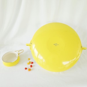 May include: A large, bright yellow round bowl with two side handles and a small matching yellow pouring vessel with a white interior. Several small, colorful oval-shaped candies are scattered between the two yellow kitchen items.