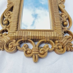 Puede incluir: Un espejo rectangular y ornamentado con un marco texturizado de color marr&oacute;n claro. El marco presenta detalles de volutas y un dise&ntilde;o acanalado. El espejo refleja un cielo azul con nubes. El estilo es vintage o antiguo.
