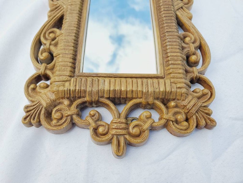 Puede incluir: Un espejo rectangular y ornamentado con un marco texturizado de color marr&oacute;n claro. El marco presenta detalles de volutas y un dise&ntilde;o acanalado. El espejo refleja un cielo azul con nubes. El estilo es vintage o antiguo.