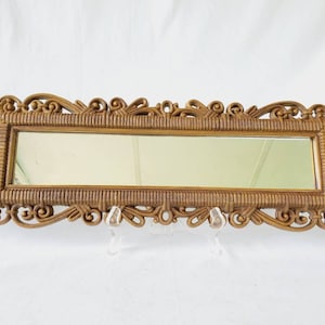 Puede incluir: Un espejo rectangular con un marco dorado ornamentado. El marco presenta una textura tejida y adornos decorativos. El espejo se exhibe sobre un soporte transparente sobre un fondo blanco.