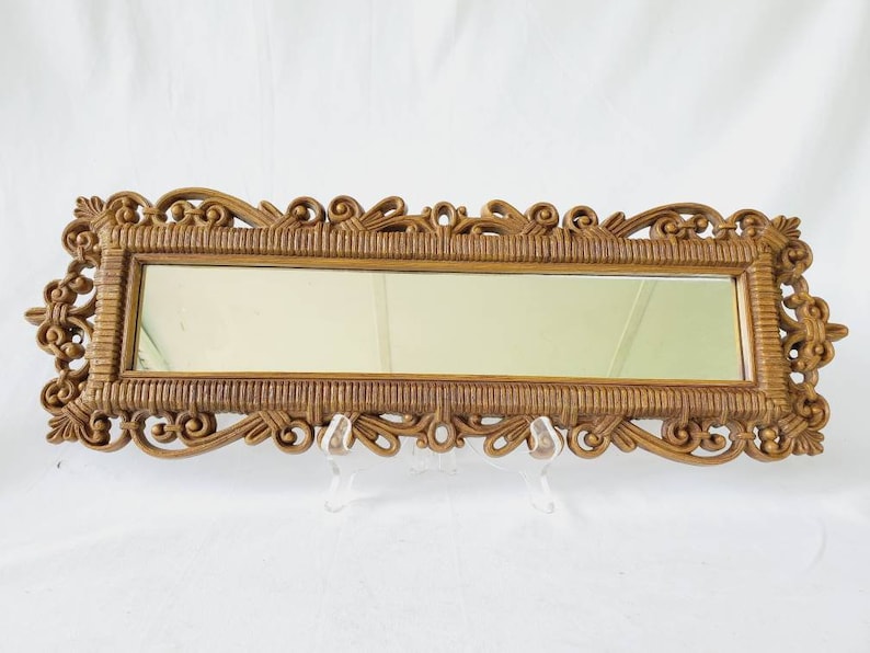 Puede incluir: Un espejo rectangular con un marco dorado ornamentado. El marco presenta una textura tejida y adornos decorativos. El espejo se exhibe sobre un soporte transparente sobre un fondo blanco.