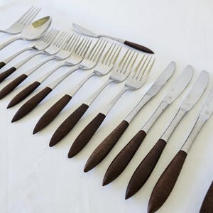 May include: A collection of stainless steel silverware with dark brown wooden handles. The set includes forks, knives, and spoons, arranged on a white surface. The silverware has a mid-century modern aesthetic.