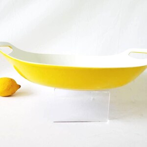 May include: A yellow serving dish with a white interior and handles. The oblong dish is on a white surface. A lemon sits beside the dish. The dish is likely for serving food.