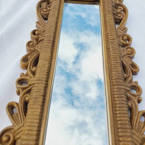 Puede incluir: Un espejo largo y rectangular con un marco dorado ornamentado. El marco presenta detalles de volutas y l&iacute;neas verticales texturizadas. El espejo refleja un cielo azul con nubes blancas y esponjosas, creando un acento decorativo de pared.