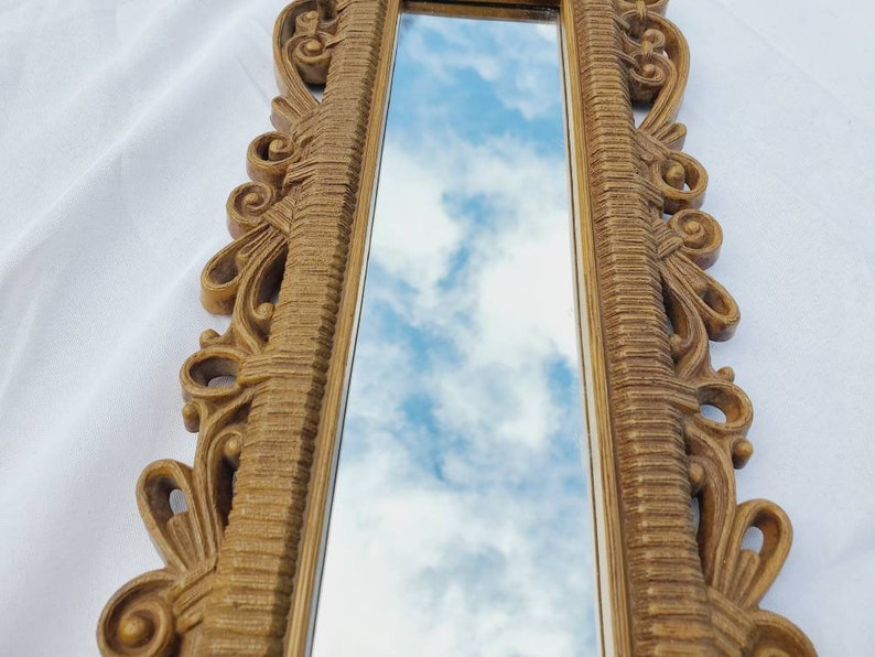 Puede incluir: Un espejo largo y rectangular con un marco dorado ornamentado. El marco presenta detalles de volutas y l&iacute;neas verticales texturizadas. El espejo refleja un cielo azul con nubes blancas y esponjosas, creando un acento decorativo de pared.