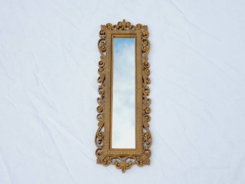 Puede incluir: Un espejo rectangular dorado y ornamentado con un marco detallado y con volutas. La superficie reflectante del espejo es larga y estrecha, reflejando un tono azul cielo. El marco presenta intrincados dise&ntilde;os florales y de volutas.