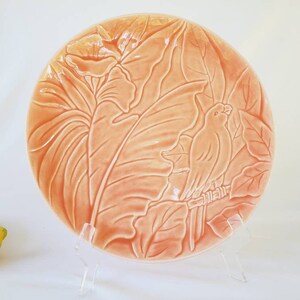 May include: A peach-coloured ceramic plate with a raised design of tropical leaves and a bird. The plate is on a white background.