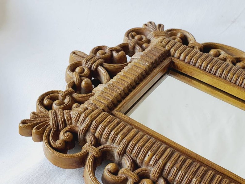Puede incluir: Primer plano de un espejo decorativo con un marco ornamentado. El marco es de color marr&oacute;n dorado c&aacute;lido y presenta intrincados adornos y detalles texturizados. La superficie del espejo refleja el entorno.