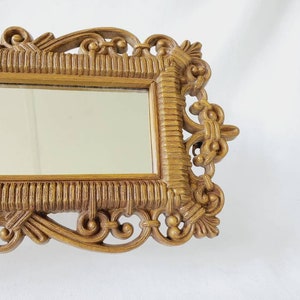 Puede incluir: Un espejo rectangular con un marco dorado ornamentado. El marco presenta intrincados adornos y un patr&oacute;n texturizado similar al tejido. La superficie del espejo refleja el entorno.