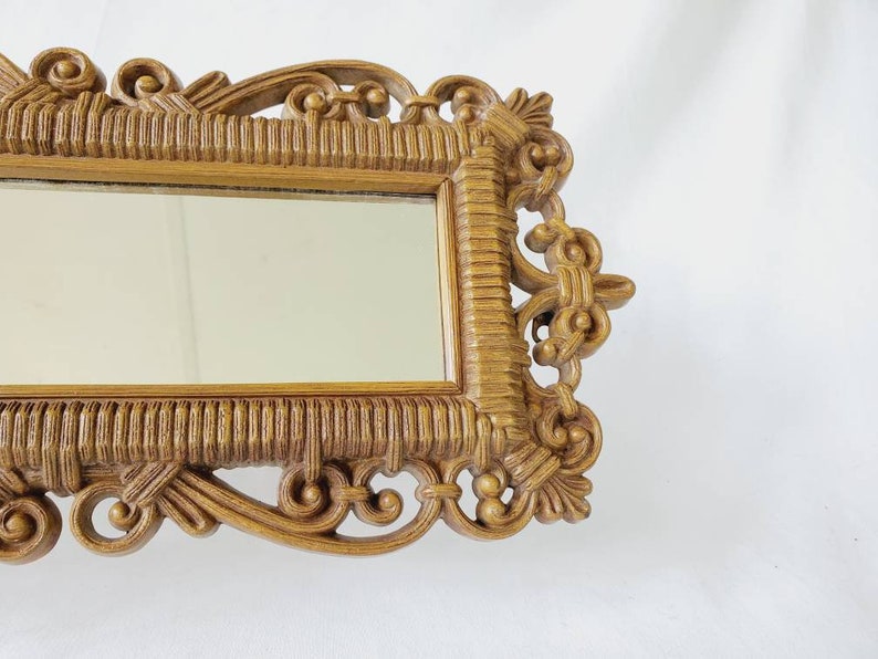 Puede incluir: Un espejo rectangular con un marco dorado ornamentado. El marco presenta intrincados adornos y un patr&oacute;n texturizado similar al tejido. La superficie del espejo refleja el entorno.