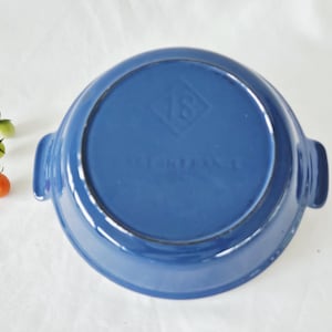 Puede incluir: Una fuente para hornear de cerámica azul cobalto con dos asas, vista desde abajo. La fuente tiene forma circular con un borde ligeramente elevado. Una ramita de tomates está a la izquierda de la fuente.