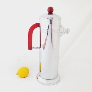 May include: A silver cocktail shaker with a red plastic handle and a red plastic top. The shaker has a cylindrical shape and a rounded base.