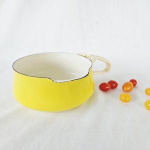 May include: A bright yellow enamel saucepan with a white interior and a cream-colored handle. The rim features a black edge with a small pouring spout. Several small red and yellow cherry tomatoes are scattered near the base of the pan.