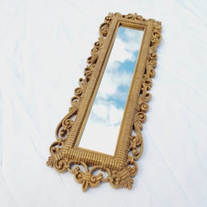 Puede incluir: Un espejo rectangular dorado y ornamentado con un marco detallado y con volutas. El espejo refleja un cielo azul con nubes blancas y esponjosas. El espejo est&aacute; en &aacute;ngulo, mostrando su dise&ntilde;o decorativo y el reflejo del cielo.