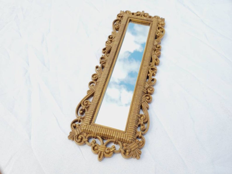 Puede incluir: Un espejo rectangular dorado y ornamentado con un marco detallado y con volutas. El espejo refleja un cielo azul con nubes blancas y esponjosas. El espejo est&aacute; en &aacute;ngulo, mostrando su dise&ntilde;o decorativo y el reflejo del cielo.