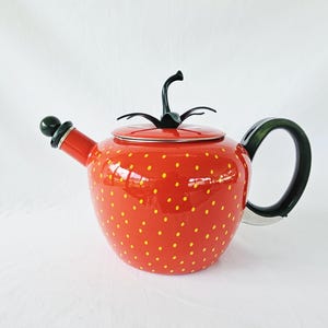 May include: A bright red teapot shaped like a strawberry with small yellow dots across the body. It features a dark green handle, a matching dark green stem and leaf lid topper, and a spout with a dark green spherical tip on a white background.
