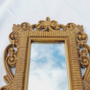 Puede incluir: Un peque&ntilde;o espejo rectangular adornado con un marco dorado. El marco presenta detalles de volutas y una superficie texturizada. El espejo refleja un cielo azul con nubes. Un objeto decorativo para la decoraci&oacute;n del hogar.