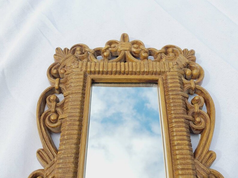 Puede incluir: Un peque&ntilde;o espejo rectangular adornado con un marco dorado. El marco presenta detalles de volutas y una superficie texturizada. El espejo refleja un cielo azul con nubes. Un objeto decorativo para la decoraci&oacute;n del hogar.