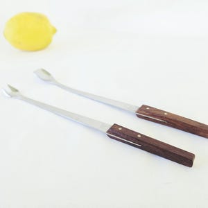 May include: Two long-handled stainless steel utensils with dark brown wooden handles secured by two small metal rivets. One utensil features a two-pronged fork tip, while the other has a small, shallow spoon-like scoop at the end.