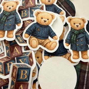 May include: A collection of die-cut stickers featuring teddy bears in various outfits. Some bears wear blue sweaters, others wear blue coats with green scarves. Wooden alphabet blocks are also present.