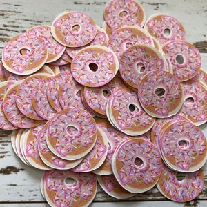 May include: A collection of circular donut-shaped cutouts. Each cutout features a pink frosted top with colorful sprinkles and a tan-colored base. The donuts are scattered across a light-colored wooden surface.