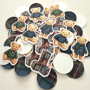 May include: A collection of paper cutouts featuring teddy bears in various outfits, including sweaters and coats. The bears are interspersed with ABC blocks and plaid and white circles. The color palette includes shades of brown, blue, green, and white.