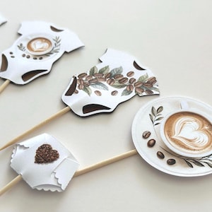 May include: White baby bodysuit-shaped cupcake toppers with coffee-themed designs. One features a latte art heart, another coffee beans, and a third a heart made of coffee beans. Each is attached to a wooden stick.