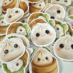 May include: A collection of cute dumpling stickers with smiling faces. The stickers feature dumplings in bamboo steamers and individual dumplings. The dumplings are white with brown eyes and pink cheeks. Some are in green lettuce.