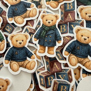 May include: A collection of die-cut stickers featuring teddy bears in various outfits. The bears are dressed in sweaters, coats, and scarves, with some sitting near alphabet blocks. The color palette includes shades of brown, blue, and green.