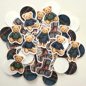May include: A collection of teddy bear stickers in various outfits, including sweaters and coats, with wooden blocks spelling "BABY". The bears are surrounded by white circles and plaid patterned circles in shades of blue, green, and brown.