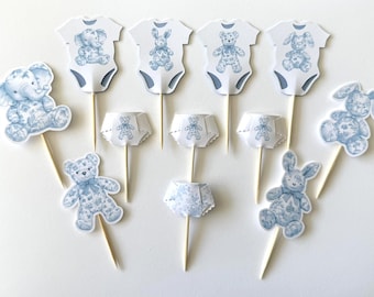 May include: A collection of baby shower cupcake toppers featuring blue and white illustrations of an elephant, teddy bears, and bunnies. The toppers include onesies, diapers, and character cutouts, all attached to wooden sticks.