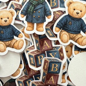May include: A collection of stickers featuring watercolor illustrations of teddy bears in various outfits, including a blue coat and a cable-knit sweater. Also included are alphabet blocks with the letters A, B, and Y, and the word "BABY".