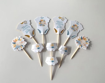 Op de afbeelding: Een verzameling cupcake toppers voor een babyshower. De toppers bevatten witte rompertjes, luiers en zonnebloemontwerpen met de tekst "Here comes the Son". De toppers zitten op houten stokjes.