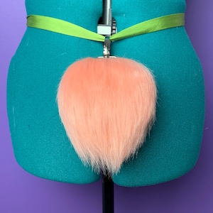 May include: A peach-colored faux fur tail attached to a green ribbon with a silver clasp. The tail is heart-shaped and fluffy. The ribbon is wrapped around a teal mannequin form, set against a purple background.