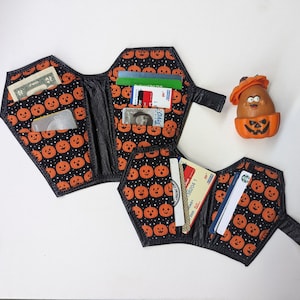 May include: A black and orange Halloween-themed wallet shaped like a coffin with a pumpkin pattern. The wallet is open and has several credit cards and cash inside.