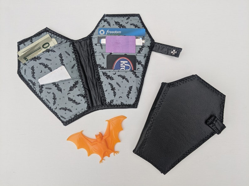 May include: A black leather wallet shaped like a coffin with a gray and black bat print interior. The wallet is open and has several cards and cash inside. There is a small orange plastic bat figurine next to the wallet.