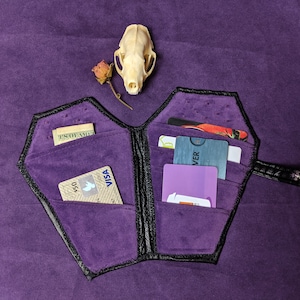 May include: A black leather wallet shaped like a coffin with a purple velvet interior. The wallet is open and has several cards and cash inside.