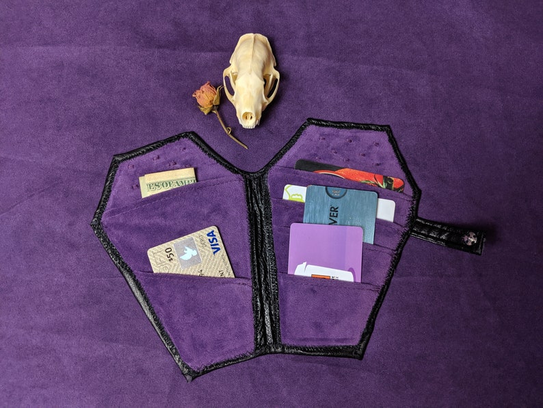 May include: A black leather wallet shaped like a coffin with a purple velvet interior. The wallet is open and has several cards and cash inside.