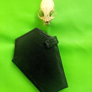May include: A black leather wallet with a stitched edge on a green background. A white animal skull is above the wallet.