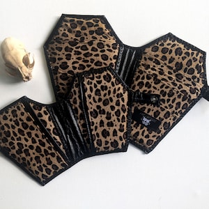 May include: A black and brown leopard print wallet shaped like a coffin. The wallet has a black flap with a snap closure and a small black strap.