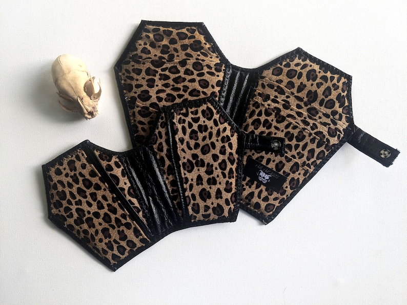 May include: A black and brown leopard print wallet shaped like a coffin. The wallet has a black flap with a snap closure and a small black strap.