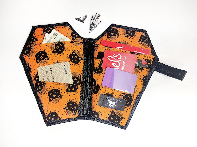 May include: A black and orange fabric wallet shaped like a coffin with a black cat pattern. The wallet is open and has several cards inside, including a Vanilla gift card and a "Happens at Fred's" gift card.