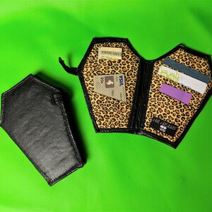 May include: A black leather coffin-shaped wallet with a leopard print lining. The wallet is open and shows several cards inside. The text "Till Death" is visible on the inside of the wallet.