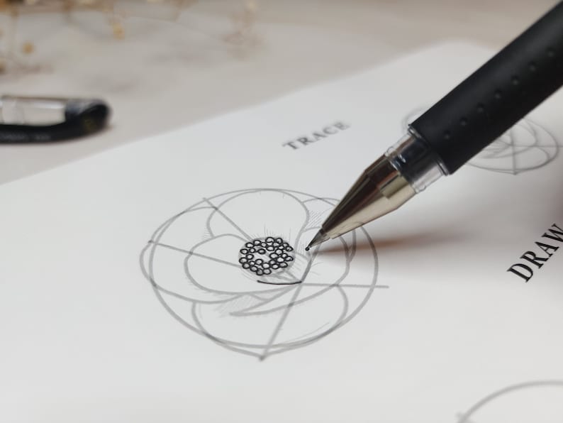 May include: A close-up of a hand using a black pen to trace a drawing of a flower on white paper. The paper has the words "TRACE" and "DRAW" written on it.