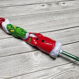 Puede incluir: Un gancho de ganchillo de temática Grinch verde y rojo con un gancho de metal verde. El Grinch lleva un gorro de Papá Noel.