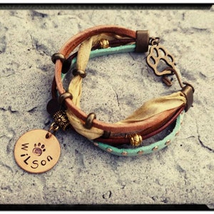 May include: A multi-layered leather bracelet with a charm that says "Wilson" and a paw print. The bracelet is decorated with gold beads and a turquoise leather strap.